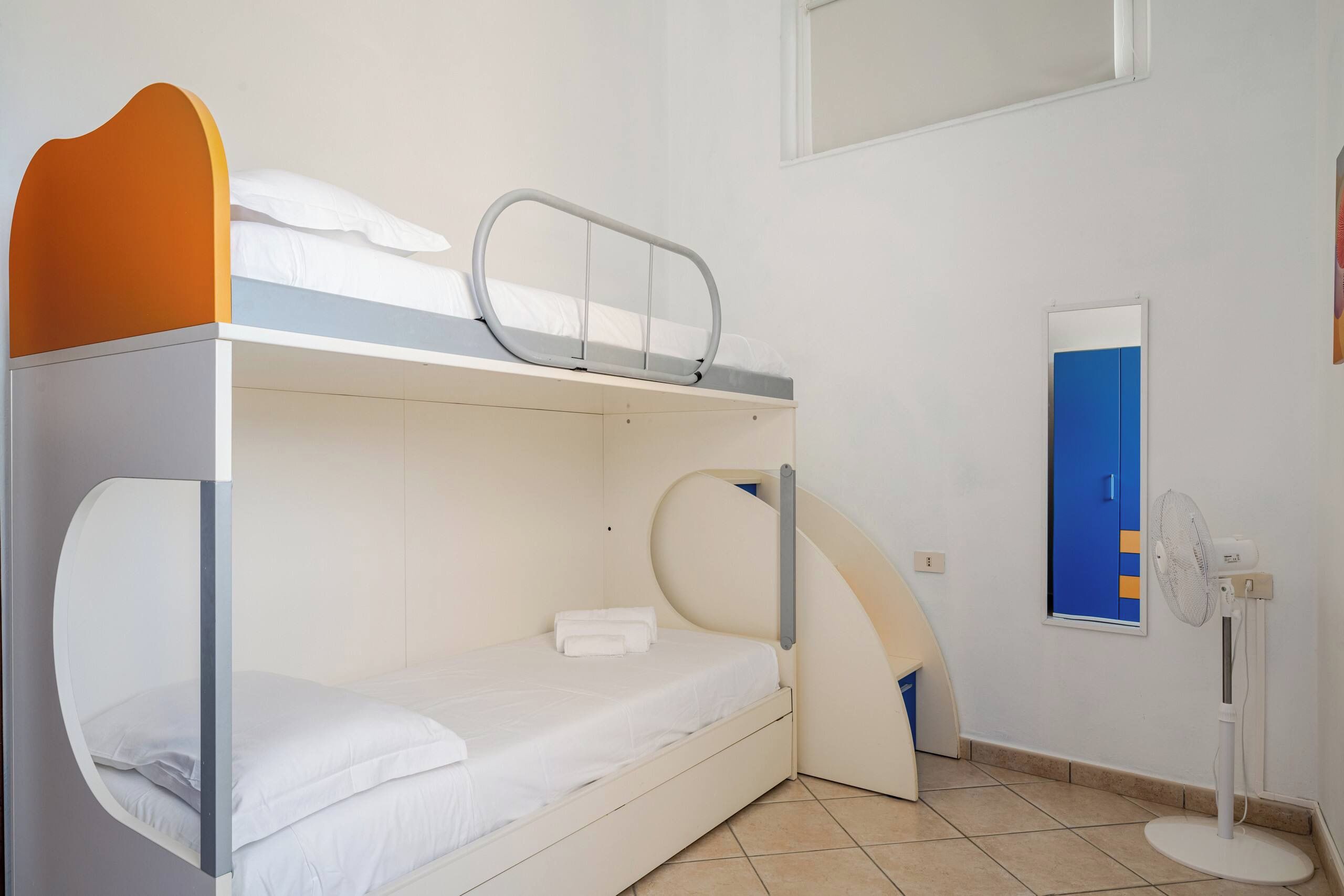 Een slaapkamer met een stapelbed, een klein bureau, een spiegel en een ventilator.