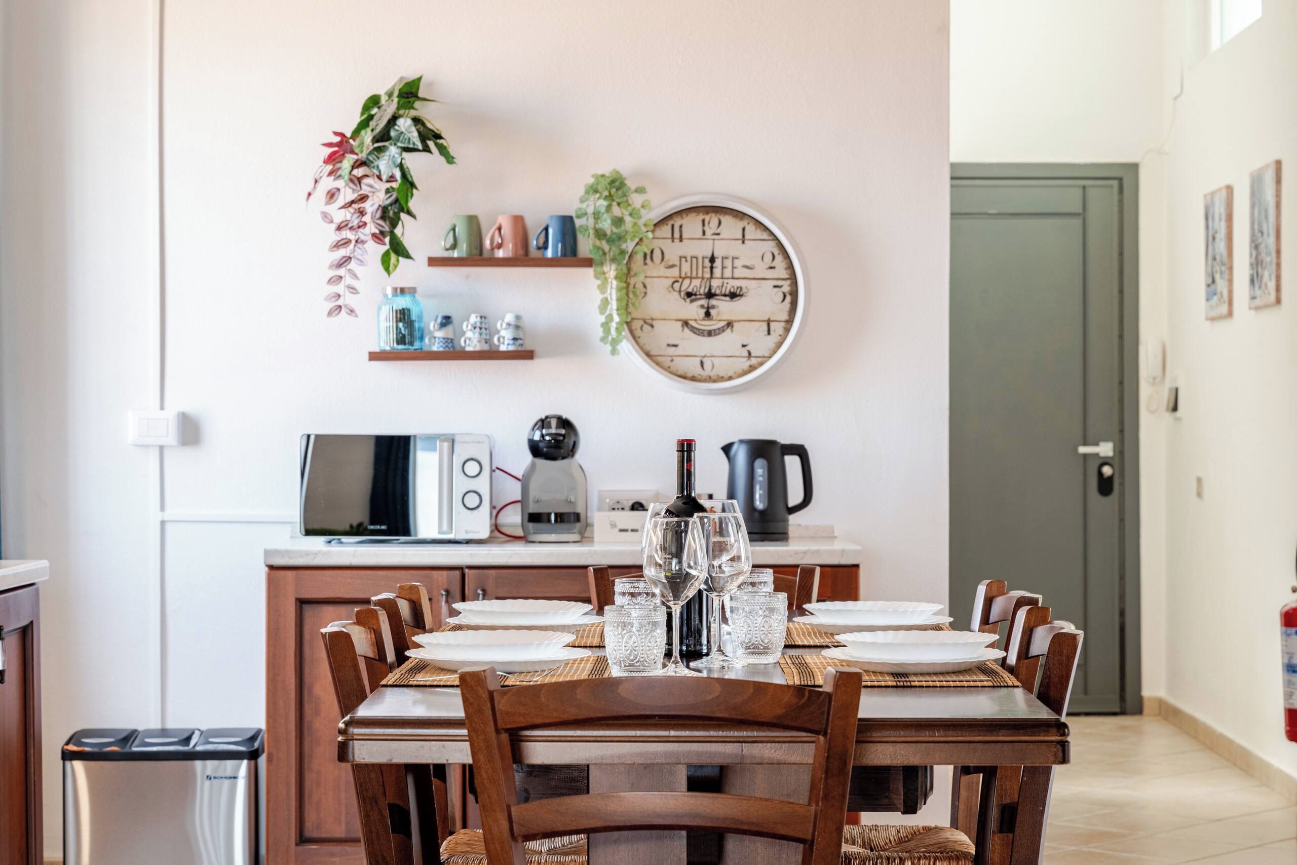 A dining area with a round table set for six, a coffee collection clock, and kitchen appliances in the background.
