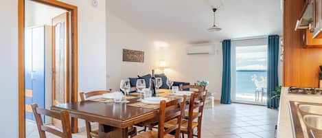 A dining area with a wooden table and chairs, a sofa, and a view of the sea through a large window.