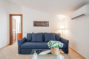 A living room with a grey sofa, a glass coffee table, a vase of flowers, a lamp, and an air conditioning unit on the wall.