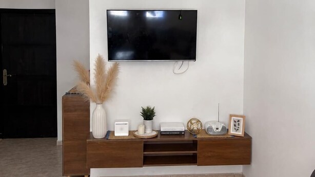 Ein Wohnzimmer mit einem an einer weißen Wand befestigten Flachbildfernseher, einer Holz-Unterhaltungseinheit darunter und einer dekorativen Pendelleuchte.