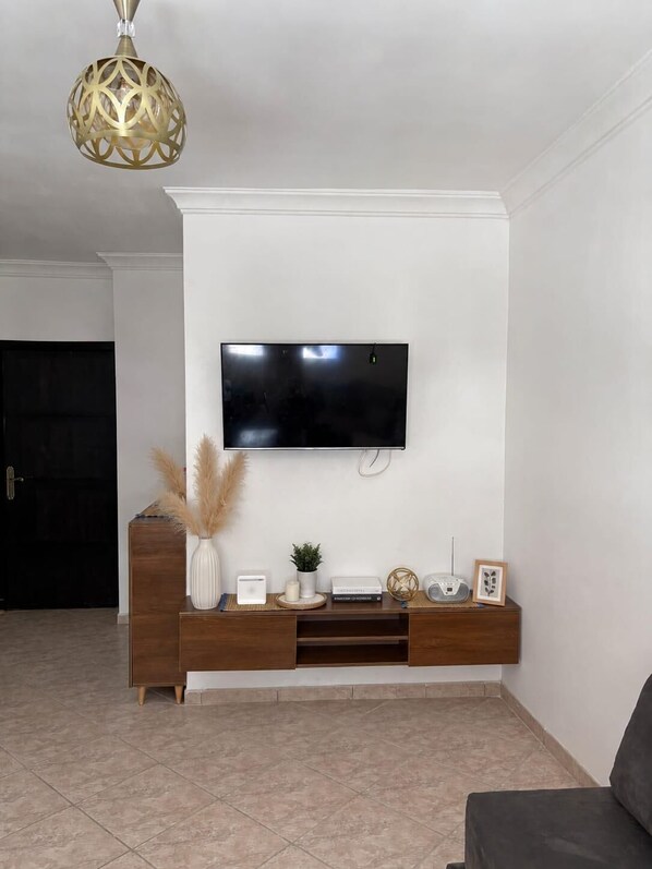 A living room with a flat-screen TV mounted on a white wall, a wooden entertainment unit below, and a decorative hanging light fixture.