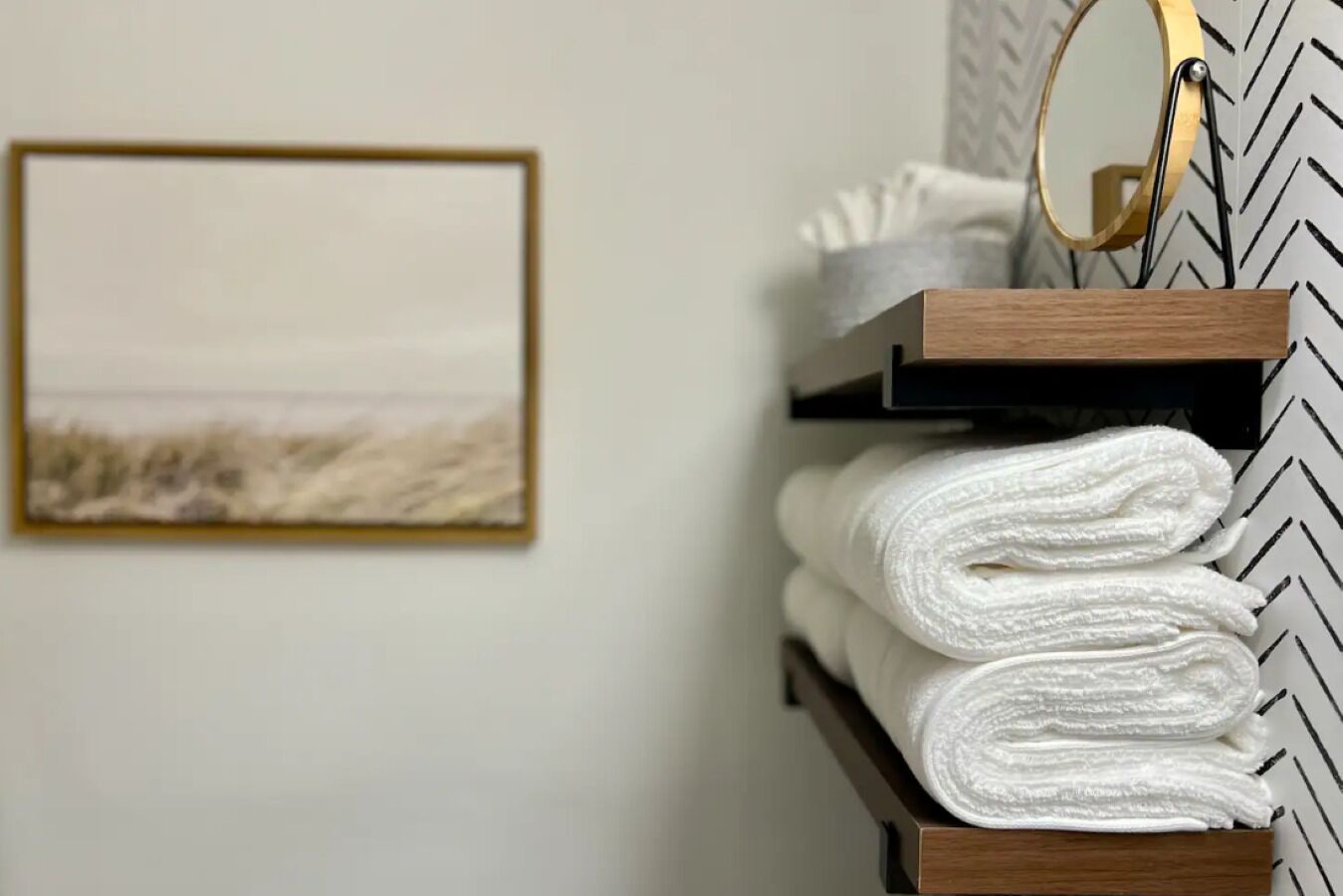 A bathroom with a wooden shelf holding white towels, a framed picture on the wall, and a mirror with a wooden frame.