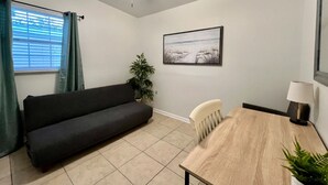 Ein Zimmer mit einem dunklen Sofa, einem Holztisch, einem Stuhl, einem Deckenventilator, einer Topfpflanze, einem gerahmten Bild an der Wand und einem Fenster mit Vorhängen.
