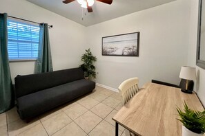 Ein Zimmer mit einem dunklen Sofa, einem Holztisch, einem Stuhl, einem Deckenventilator, einer Topfpflanze, einem gerahmten Bild an der Wand und einem Fenster mit Vorhängen.