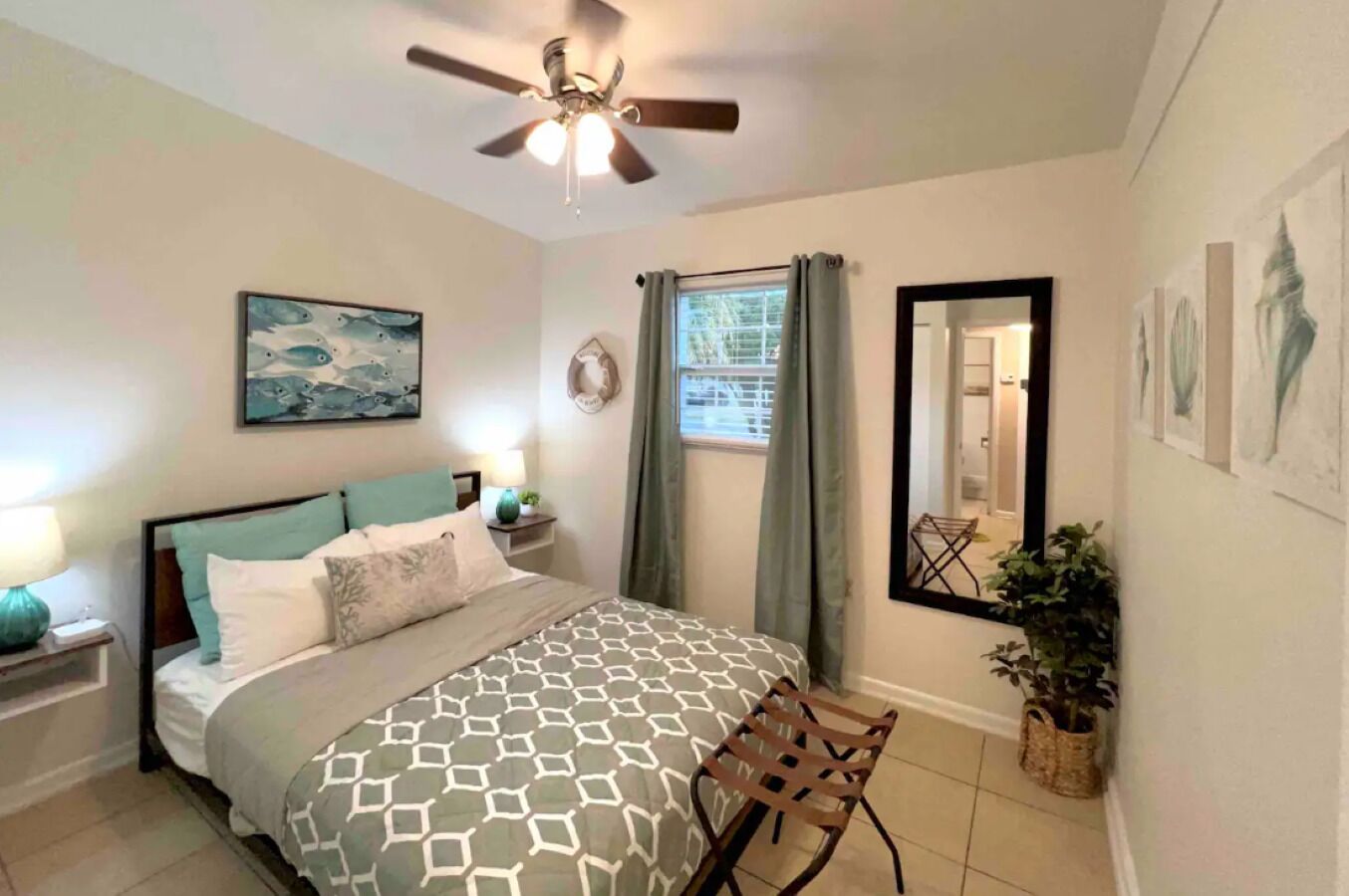 Une chambre à coucher avec un lit, un ventilateur de plafond, une fenêtre avec des rideaux, un miroir et une chaise.