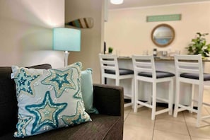 Ein Wohnzimmer mit einem dunkelbraunen Sofa, einem Kissen mit blaugrün-weißem Sternenmuster, einer weißen Lampe und einem Essbereich mit weißen Stühlen und einem Tisch.