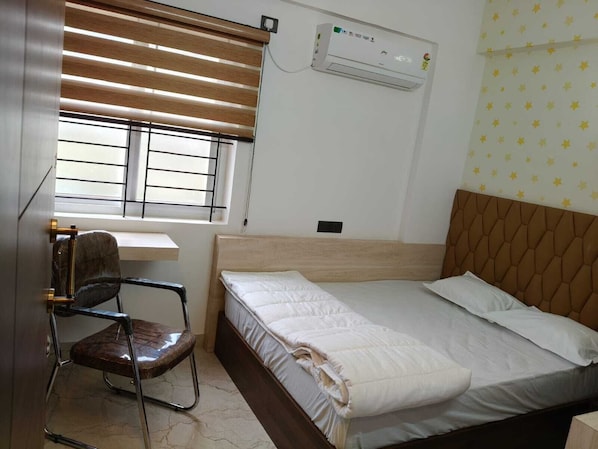 A bedroom with a bed, a desk, a chair, an air conditioning unit, and a window with blinds.