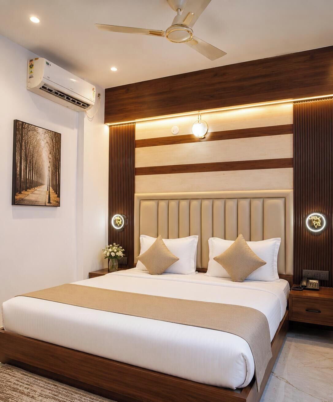 Habitación de hotel con una cama grande, ventilador de techo, aire acondicionado, mesita de noche con lámpara y una obra de arte colgada en la pared.