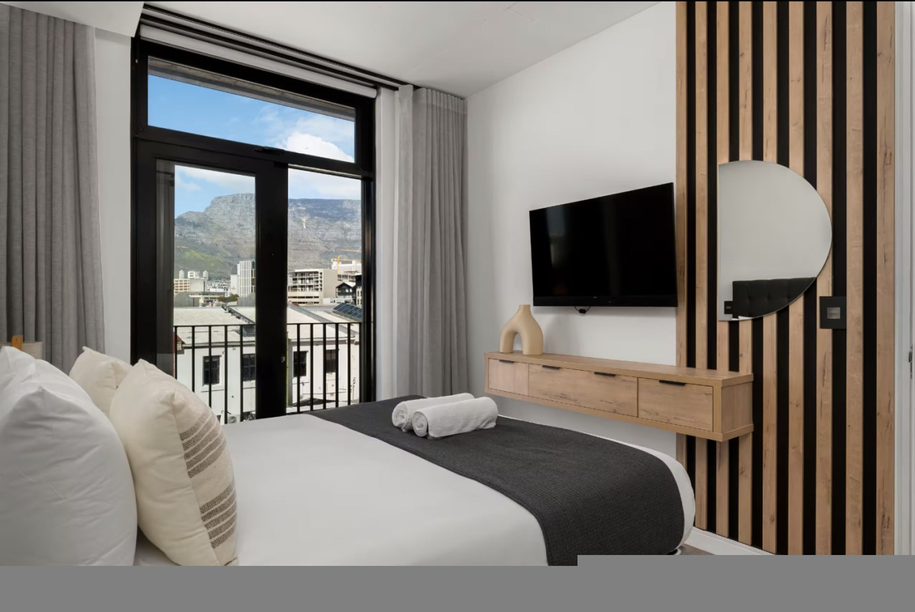 Habitación de hotel con cama, televisor montado en la pared, mueble de madera y vista de la ciudad a través de ventanas grandes.