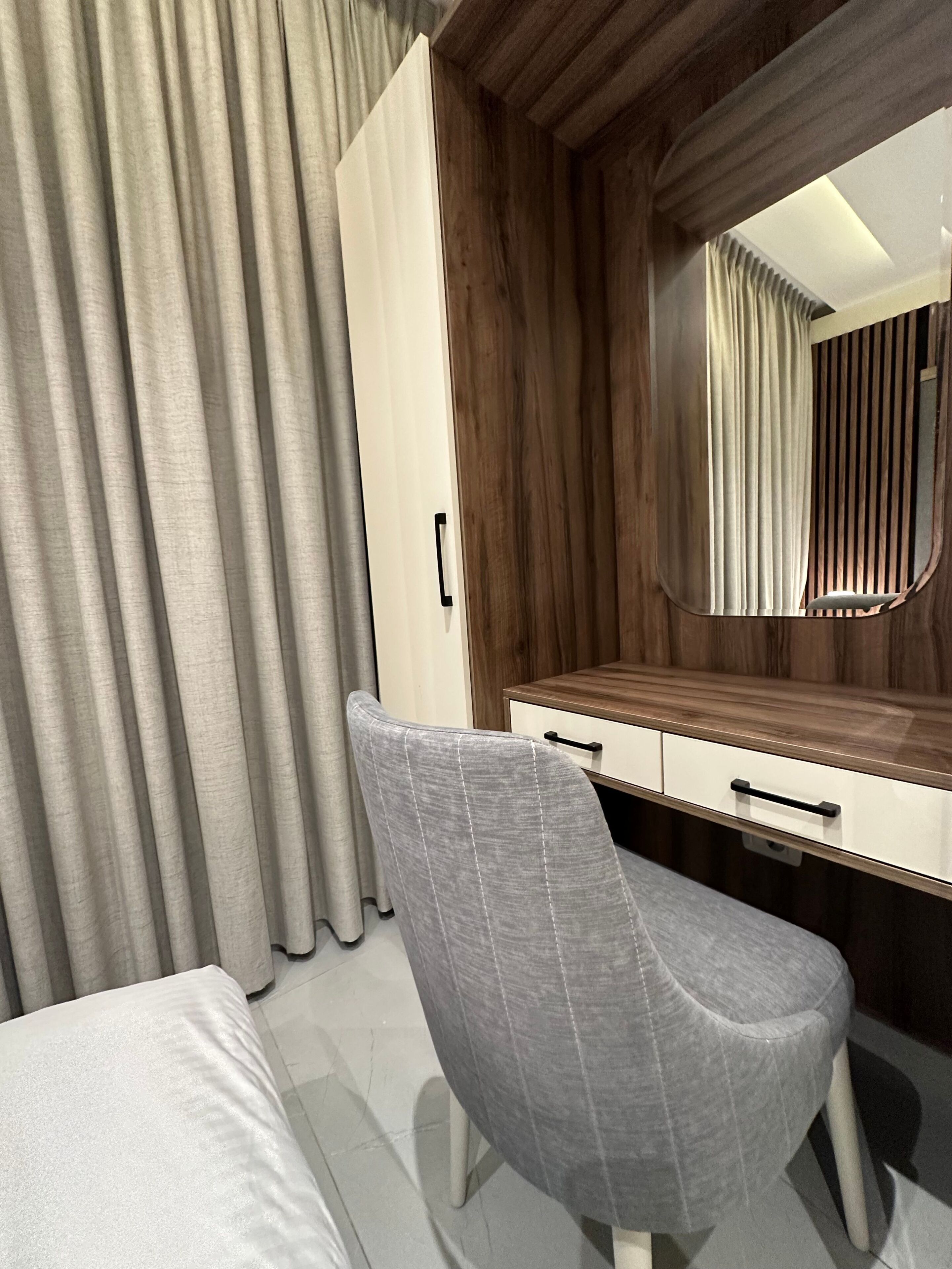Une chambre à coucher moderne avec un fauteuil à revêtement gris, une coiffeuse en bois avec un miroir et un lit doté d’une tête de lit blanche.