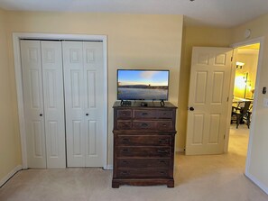 Ein Zimmer mit Fernseher auf einer Holzkommode, einer geschlossenen weißen Tür und einem Flur, der in ein weiteres Zimmer führt.