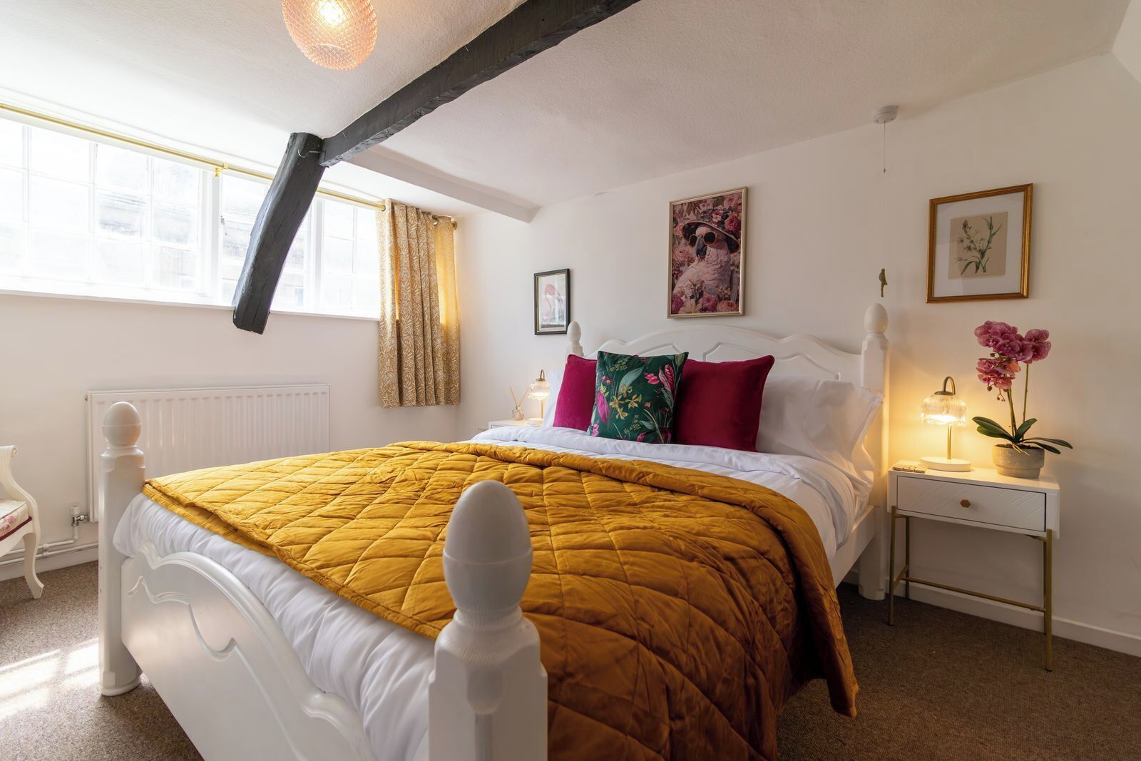 A bedroom with a white bed, a mustard-colored quilt, a nightstand with a lamp, and a window with curtains.