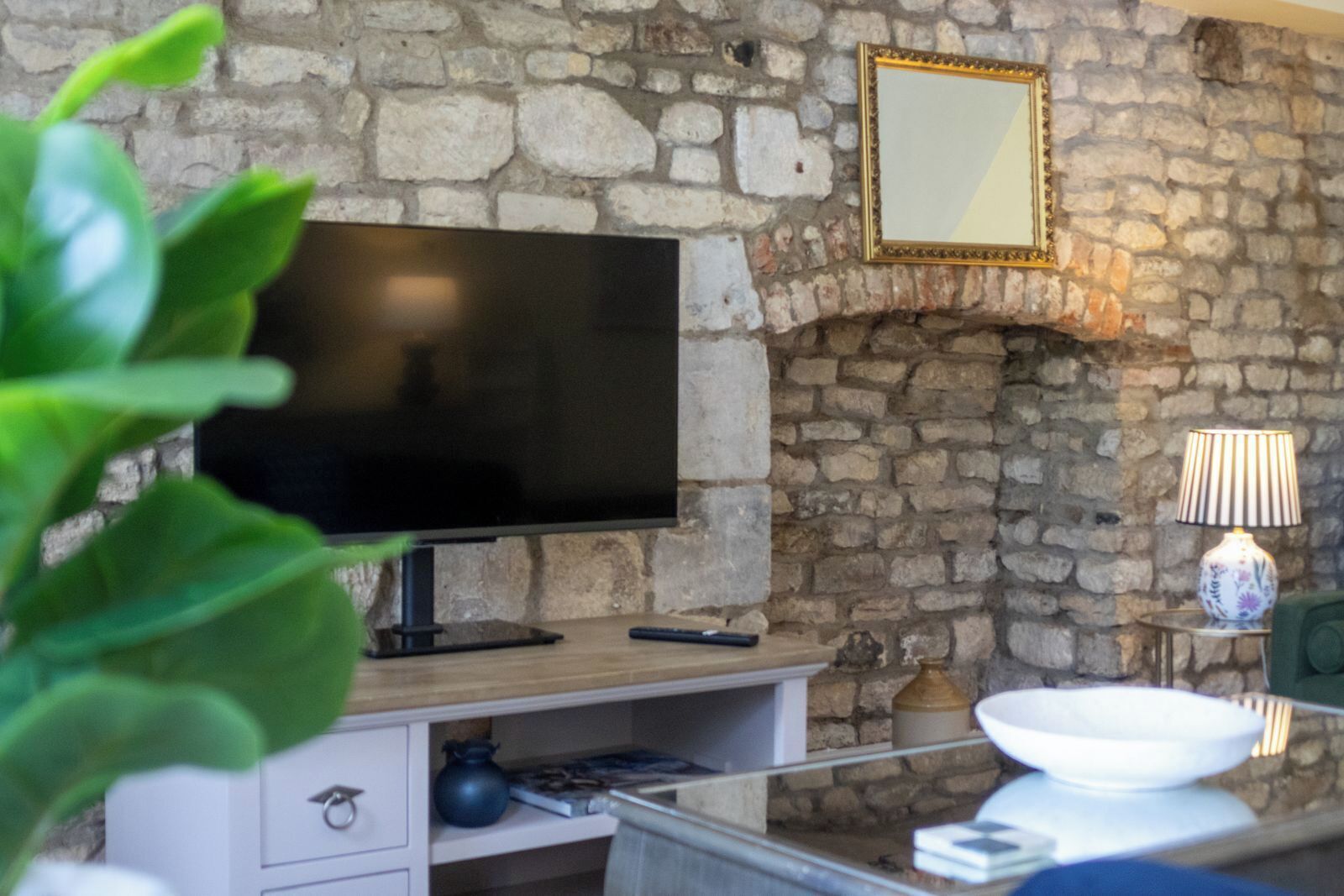 A stone wall with a flat-screen TV, a small table with a lamp, and a bowl on a shelf.