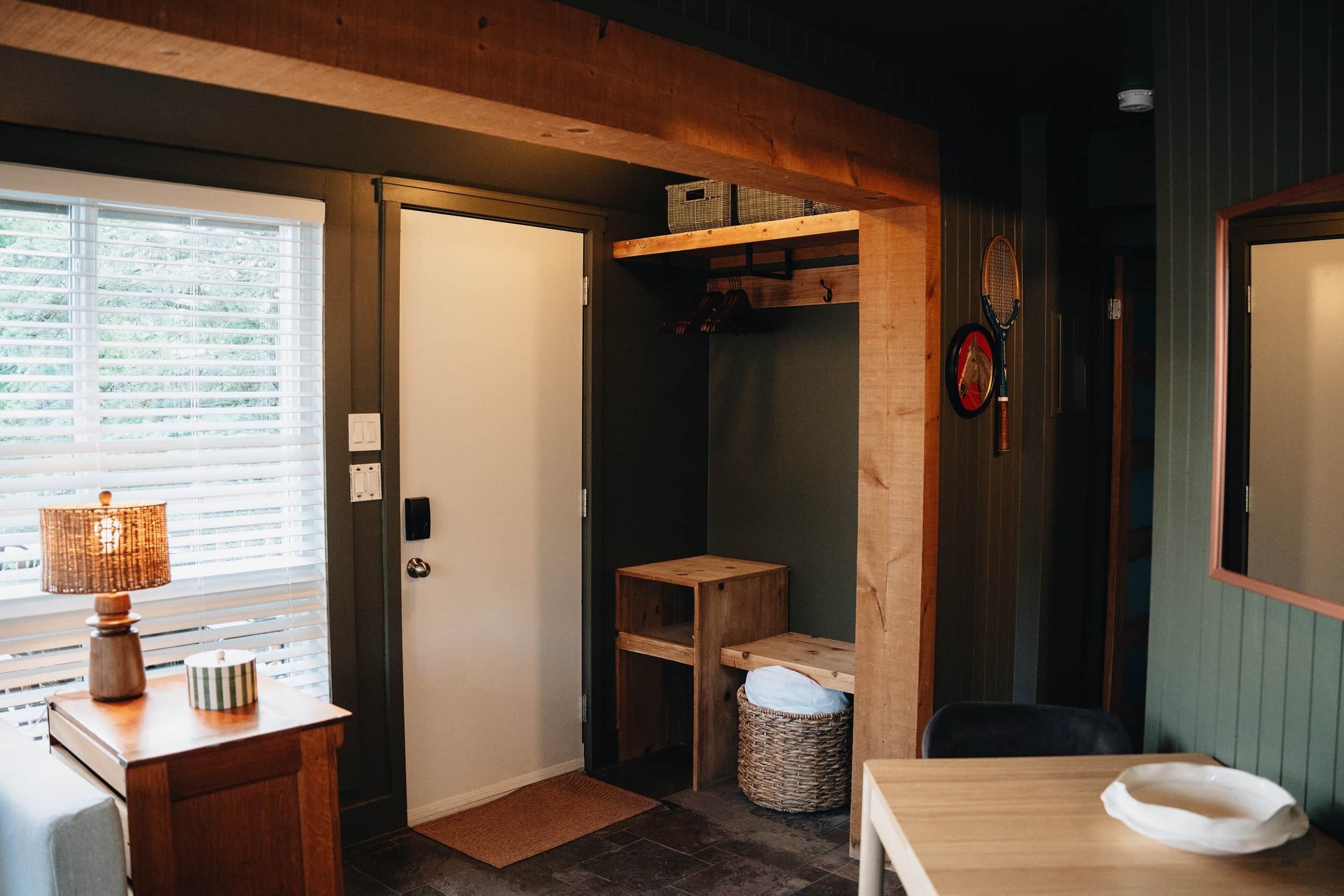 A room with a wooden door, a small table with a lamp, a wicker basket, and a wooden shelf with boxes.