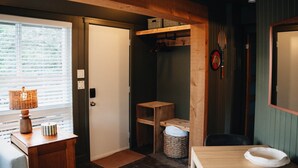 Una habitación con puerta de madera, una mesita con una lámpara, una cesta de mimbre y un estante de madera con cajas.