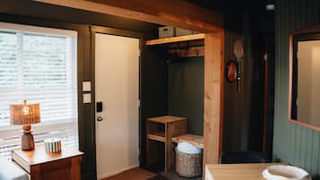 A room with a wooden door, a small table with a lamp, a wicker basket, and a wooden shelf with boxes.