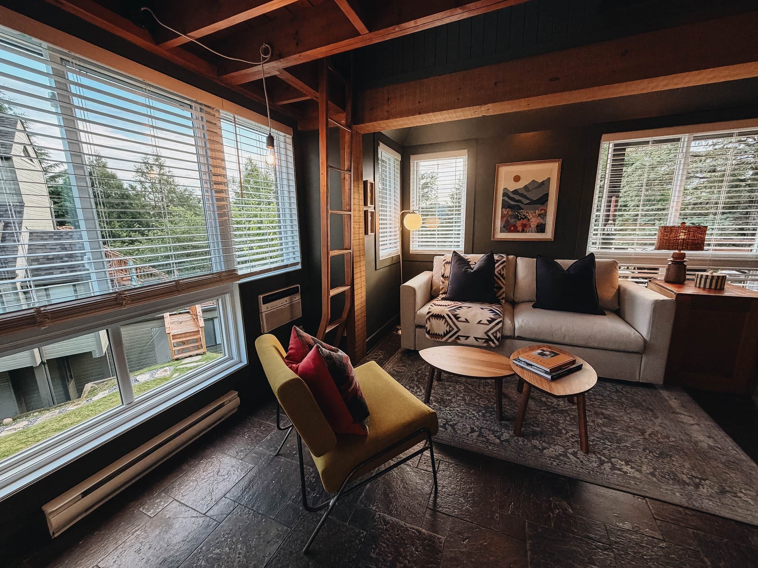 Un salón acogedor con sofá, sillón y mesa de centro, ventanas amplias con persianas y una chimenea.