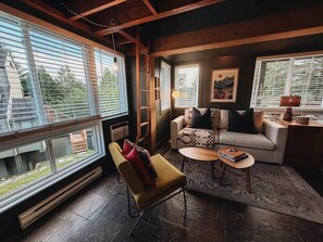Ein gemütliches Wohnzimmer mit einer Couch, einem Sessel und einem Couchtisch, großen Fenstern mit Jalousien und einem Kamin.