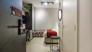 A compact hotel room with a bed, a small table, a TV, a minibar, and a door with a number sign. - StayHome VN Turiassu | Gym & Pool by Allianz Parque (São Paulo)