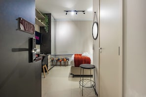 A compact hotel room with a bed, a small table, a TV, a minibar, and a door with a number sign. - StayHome VN Turiassu | Gym & Pool by Allianz Parque (São Paulo)