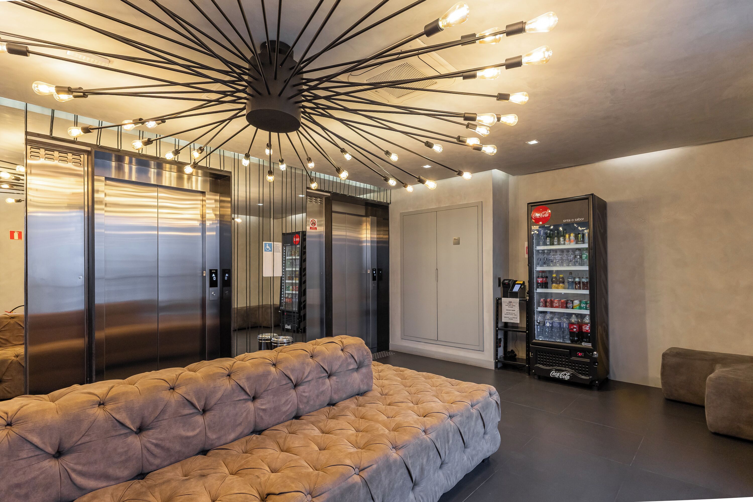 A lobby with a tufted sofa, elevators, a vending machine, and a modern ceiling light fixture.