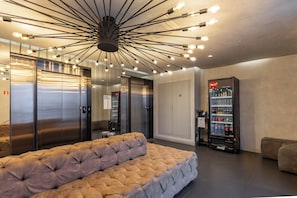 A lobby with a tufted sofa, elevators, a vending machine, and a modern ceiling light fixture. - StayHome VN Turiassu | Gym & Pool by Allianz Parque (São Paulo)