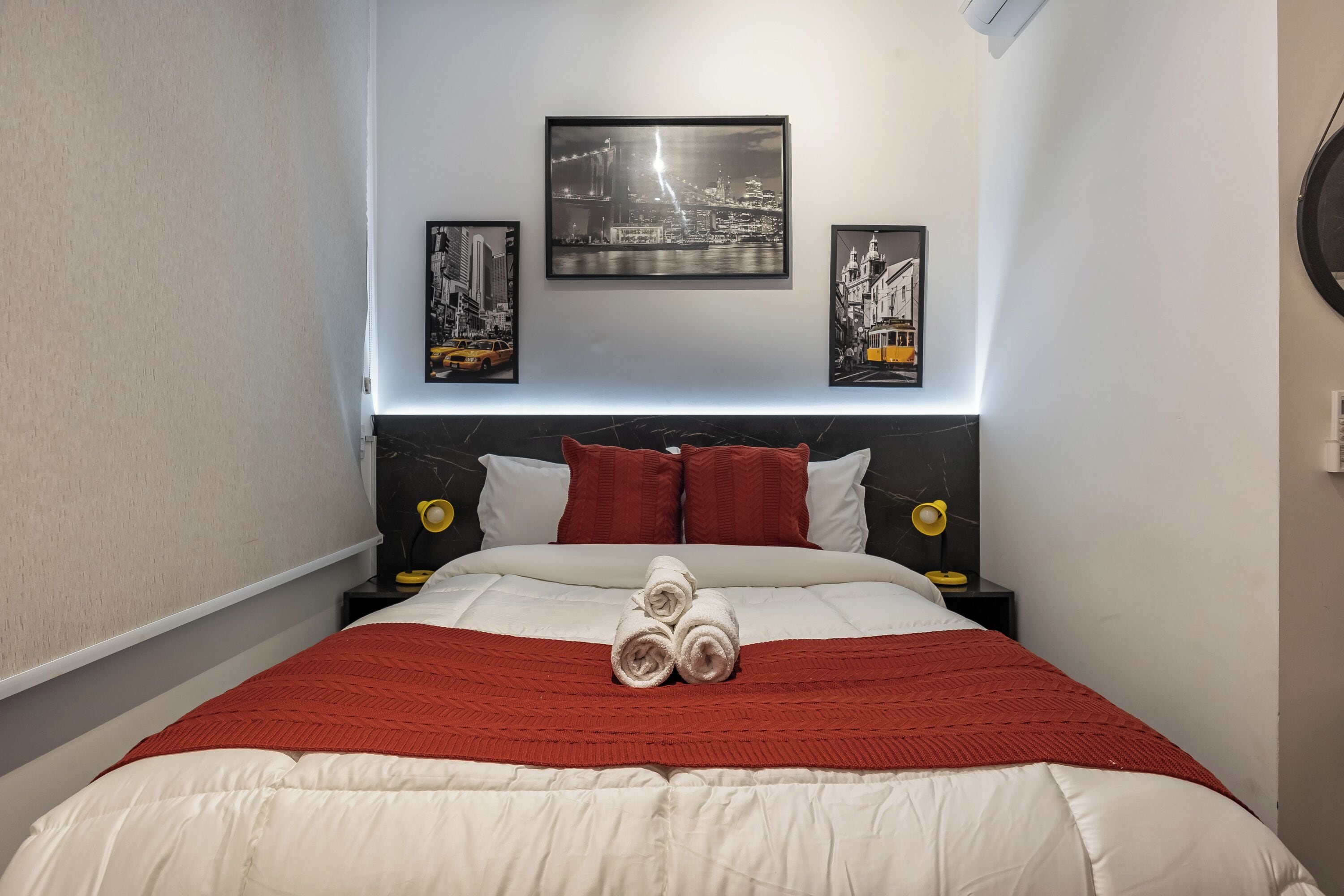 Uma cama bem arrumada com um cobertor vermelho, uma obra de arte com tema de paisagem urbana acima e uma cabeceira com textura semelhante ao mármore.