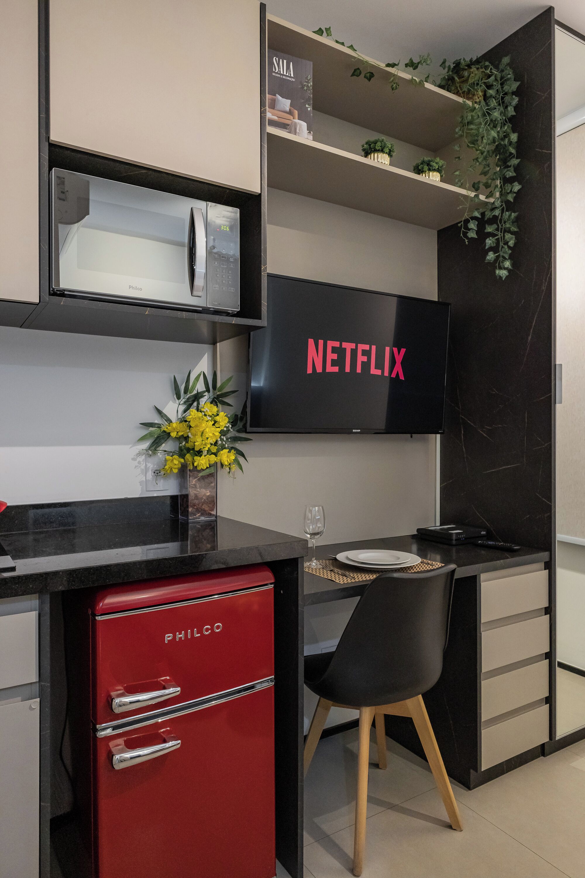 Ett modernt kök med ett rött kylskåp från Philco, en svart bänkskiva, ett matbord med en stol och en väggmonterad tv som visar Netflix-logotypen.