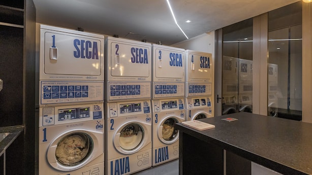 A row of four washing machines with numbered labels, a control panel, and a counter with a menu. - StayHome VN Turiassu | Gym & Pool by Allianz Parque (São Paulo)