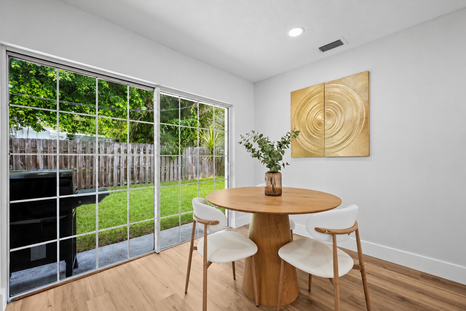 A modern dining area with a round wooden table, four white chairs, and a large window with a view of a backyard.