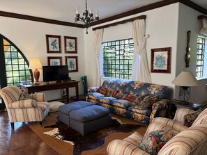 Ein Wohnzimmer mit Sofa, Hocker, Sesseln, Fernseher und Bildern an den Wänden.