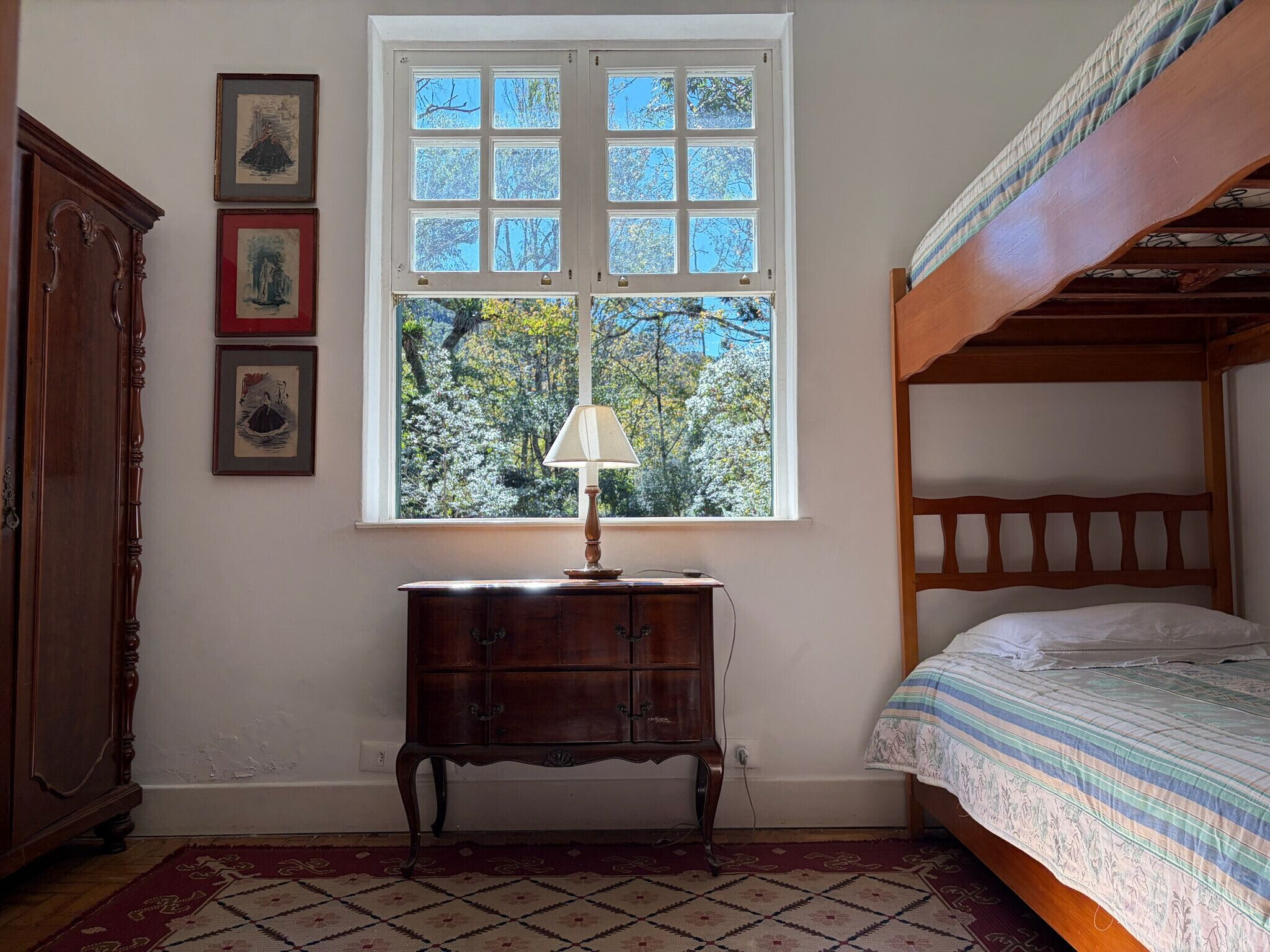 Ein Zimmer mit einem hölzernen Bett, einem Fenster mit Blick auf Bäume, einer kleinen Holzkommode und einer Lampe.