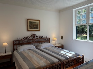 A bedroom with a wooden bed, two bedside tables, a nightstand, a framed picture on the wall, and a window with a view of greenery. - Historic Mansion with Fireplace in Campos do Jordão (Campos do Jordão)