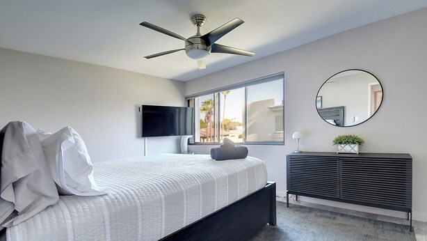 Ein modernes Schlafzimmer mit Bett, Deckenventilator, Fernseher, Fenster, Spiegel und einer Ablagekommode.