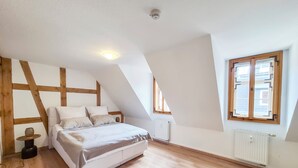 Une chambre avec un plafond en pente, un lit recouvert de linge de lit blanc, un plancher en bois et une fenêtre dotée d’un cadre géométrique.