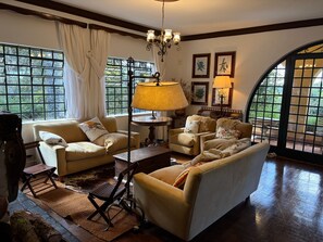 A cozy living room with a sofa, armchairs, a coffee table, and a lamp. There are windows with curtains, a chandelier, and framed artwork on the wall. - Historic Mansion With Fireplace in Campos do Jord o (Campos do Jordão)