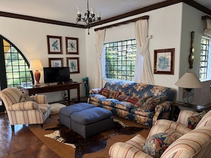 A living room with a sofa, ottoman, armchair, TV, and a chandelier. - Historic Mansion With Fireplace in Campos do Jord o (Campos do Jordão)