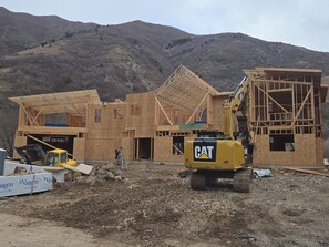 Eine Baustelle mit einem teilweise errichteten Holzbau, einem gelben CAT-Bagger und einem Arbeiter auf einer Leiter.