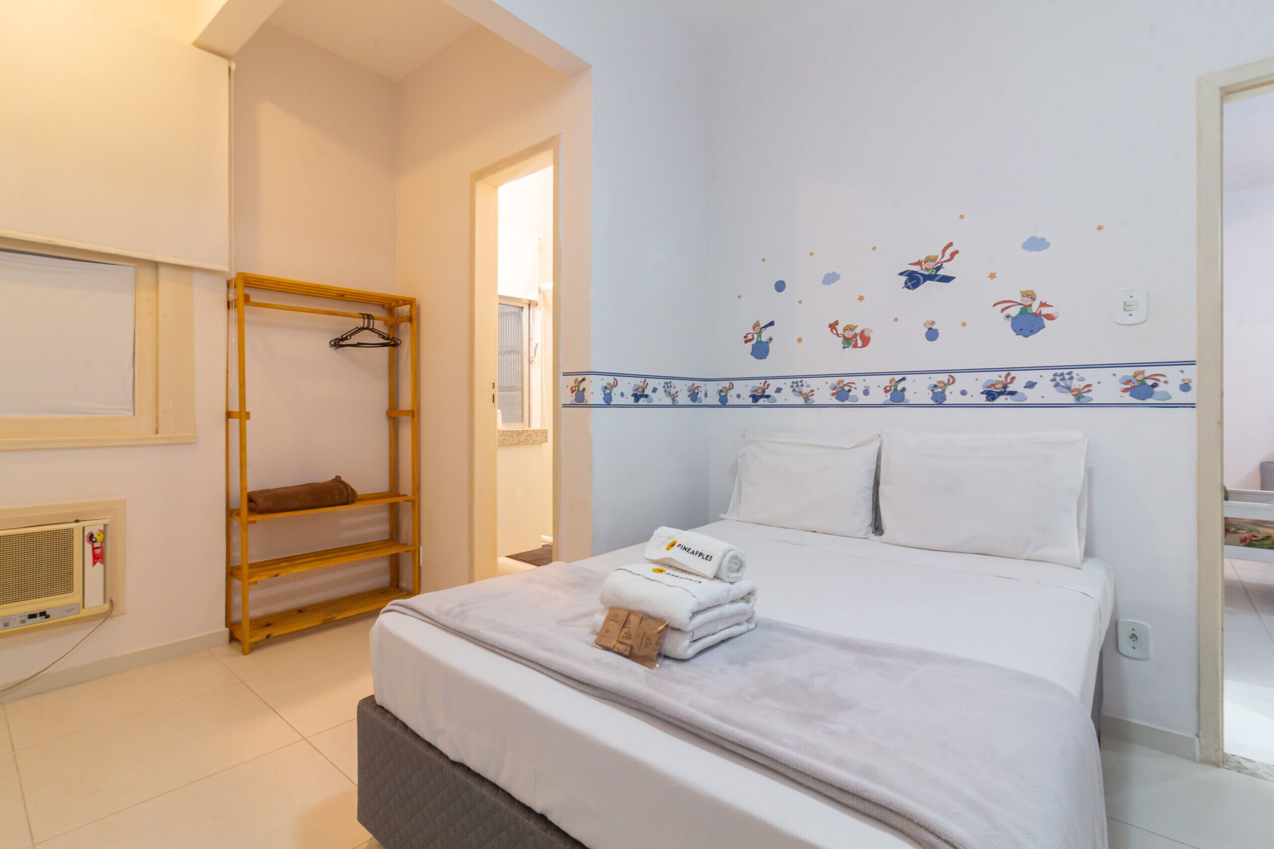 A hotel room with a bed, a wooden shelf, a wall-mounted heater, and a window.