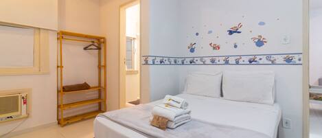 A hotel room with a bed, a wooden shelf, a wall-mounted heater, and a window.