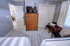 Quarto com cômoda, televisão em cima, duas camas, uma cadeira e porta que dá para outro espaço.