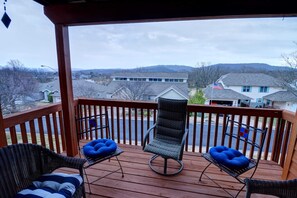 Um deck de madeira com duas cadeiras e uma mesa, com vista para uma área residencial com casas e colinas.