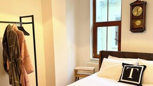 A hotel room with a bed, a wooden nightstand, a window with a view, a coat rack with hanging clothes, and a wall clock. - Redecorated Flat With Huge Library 100mbps (Istanbul)