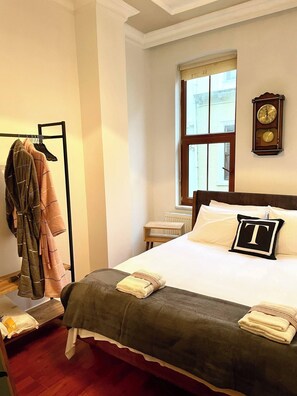 A hotel room with a bed, a wooden nightstand, a window with a view, a coat rack with hanging clothes, and a wall clock. - Redecorated Flat With Huge Library 100mbps (Istanbul)