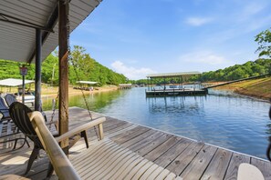 A wooden deck with a swing chair and a bench overlooking a calm river with a dock and covered seating area. - Lake Hartwell Home + Cottage, Private Dock, Hot Tub, Game Room with pool table! (Hartwell)