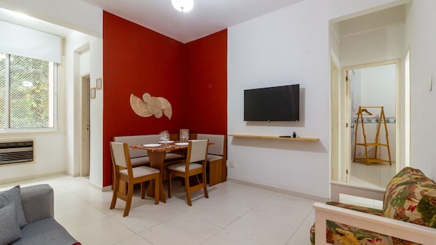 Un comedor con una pared de acento roja, un televisor montado sobre un estante de madera, y una sala de estar con un sofá y un sillón estampado.