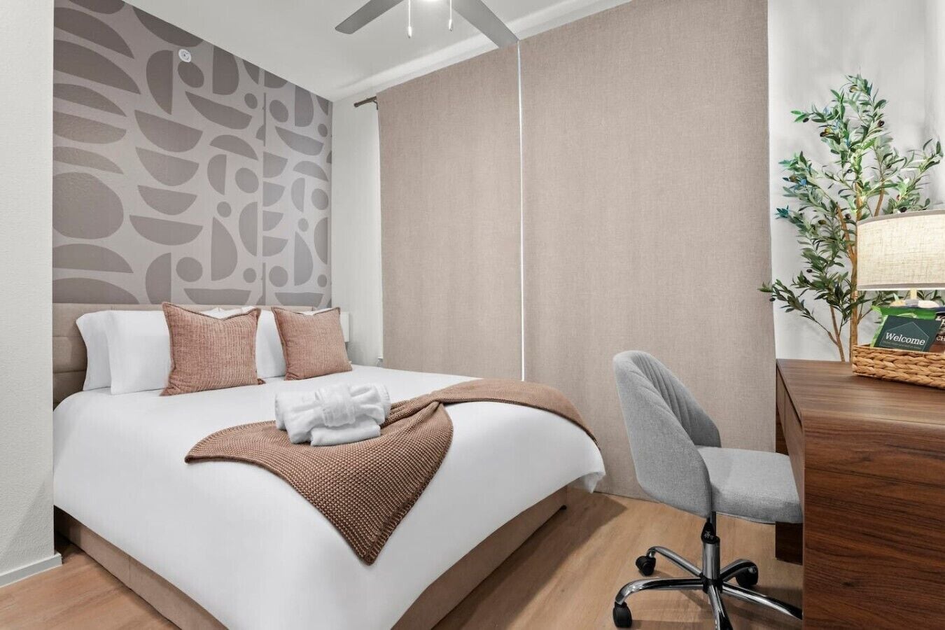 Habitación de hotel con cama, escritorio, silla, planta y tarjeta de bienvenida.