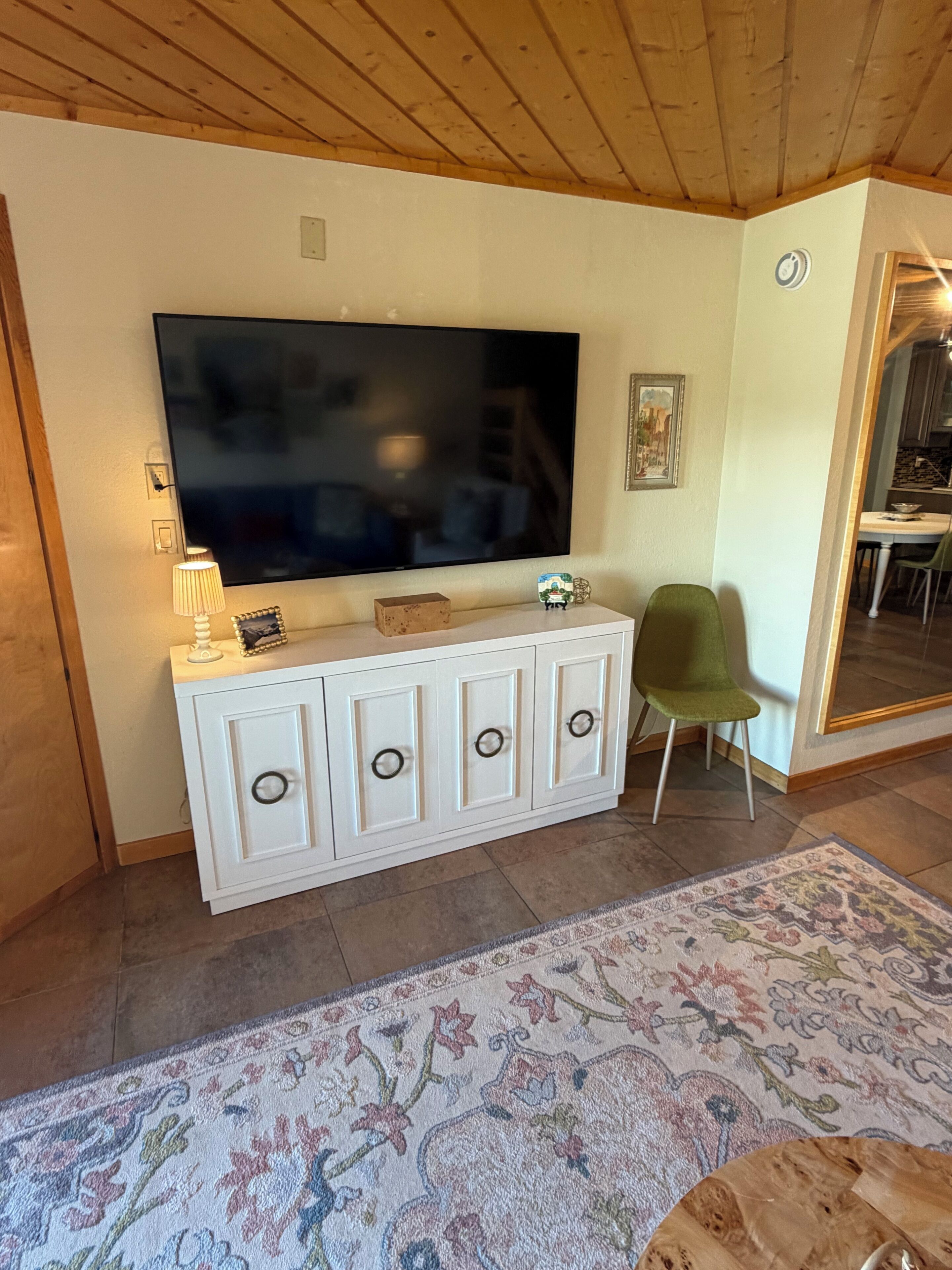 Un salon avec un téléviseur à écran plat fixé au mur, un meuble blanc avec des tiroirs, un fauteuil vert et un tapis à motifs sur le sol.
