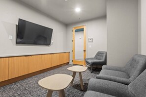 Ein Zimmer mit einem an der Wand montierten Flachbildfernseher, einem Holzschrank darunter, einem grauen Sofa, einem grauen Sessel und einem kleinen runden Tisch.